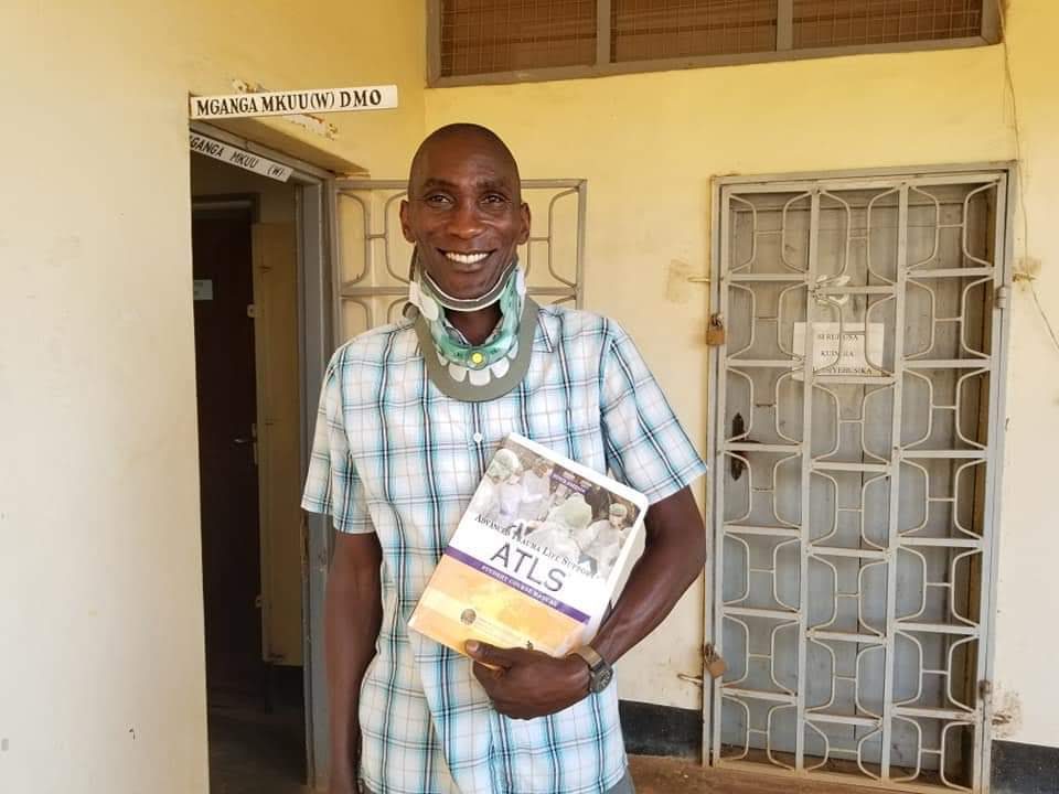Matayo Oweya smiling outside the District Medical Officer's office in Mpwapwa, holding an ATLS trauma manual and wearing a cervical collar from a training session